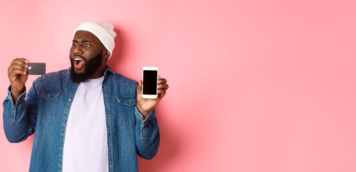 Online shopping. Shocked Black man showing mobile phone screen, looking startled at credit card, standing in hipster clothes against pink background
