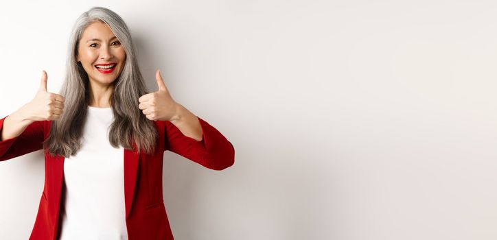 Portrait of senior asian woman showing thumb-up in approval, wearing red blazer for office work, recommend company, standing over white background