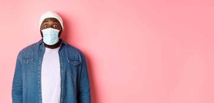 Covid-19, lifestyle and quarantine concept. Image of impressed african-american man in face mask staring at camera, gasping surprised, standing over pink background