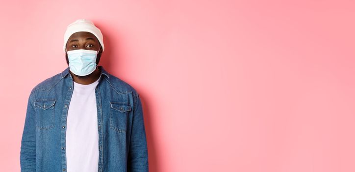 Covid-19, lifestyle and quarantine concept. Happy Black guy in beanie and face mask smiling with eyes, standing over pink background