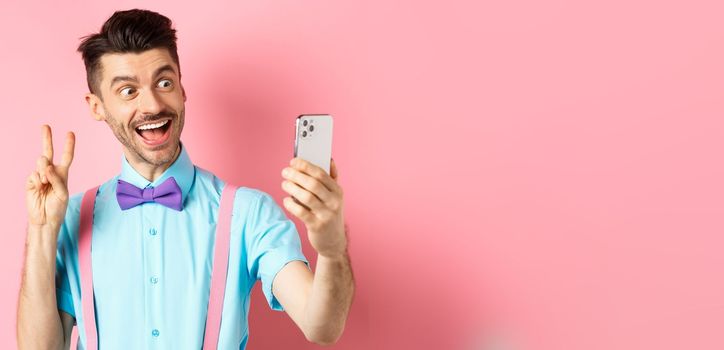 Technology concept. Funny man with moustache and bow-tie taking selfie on smartphone, showing peace sign and smiling at mobile camera, pink background