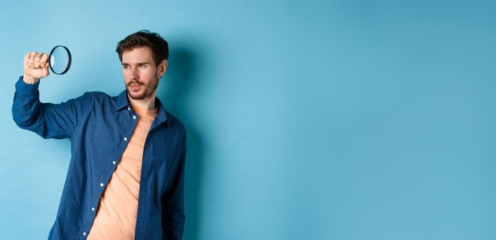 Image of young man searching for something, looking at empty space with magnifying glass, standing on blue background