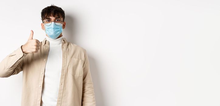 Covid-19, health and real people concept. Satisfied guy in sterile face mask and glasses, showing thumb up in approval, give positive feedback, standing on white background
