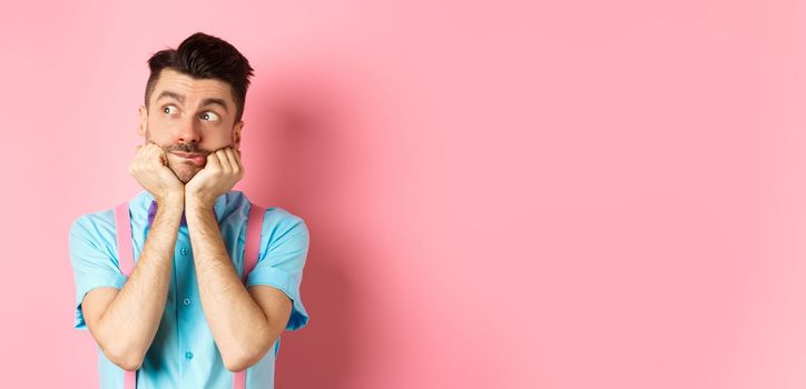 Image of silly and cute young man in suspenders, dreaming of something, looking left with curious face, standing over pink background