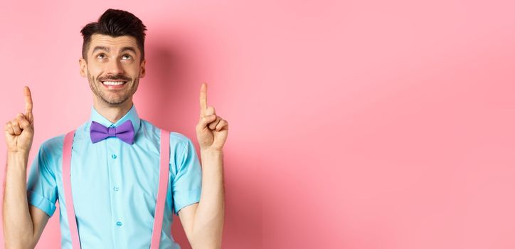 Happy smiling man in bow-tie pointing, looking up, showing something cool, standing on pink background