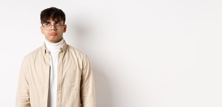 Image of sad handsome guy feeling disappointed, pouting lips and looking at empty space distressed, standing on white background in stylish clothes and eyewear