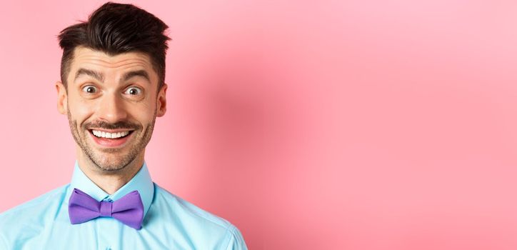Close-up of cheerful caucasian man smiling happy, looking at something interesting, standing in bow-tie over pink background