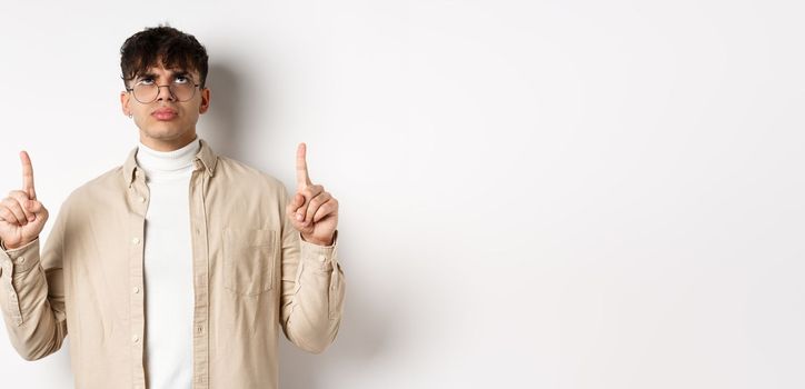 Image of annoyed or offended young man looking, pointing fingers up bothered, frowning disappointed, standing on white background