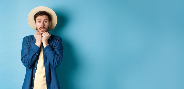 Scared young tourist trembling from fear, looking at something scary on summer vacation, wearing straw hat, standing on blue background