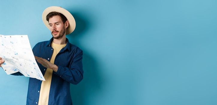 Image of handsome caucasian man looking at travel map, studying route during summer vacation, standing in straw hat on blue background
