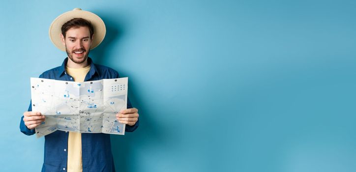 Cheerful tourist in summer hat planning travel route on vacation, looking at sighseeing map and smiling excited, standing on blue background