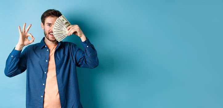 Cheerful young man showing OK sign and cover half of face with dollars, standing on blue background