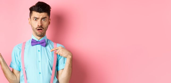 Shocked and confused guy pointing at himself with disbelief, being accused, standing on pink background in bow-tie and suspenders
