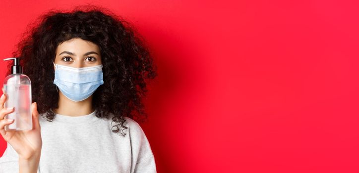 Covid-19, pandemic and quarantine concept. Young woman in medical mask showing bottle of hand sanitizer, demonstrate antiseptic, red background