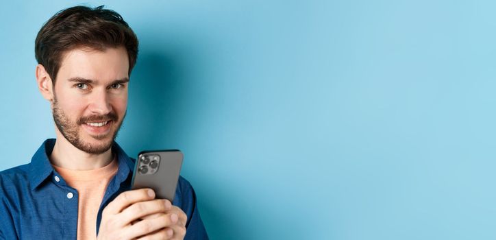 Close-up of attractive caucasian man holding mobile phone and smiling at camera, standing in casual outfit on blue background