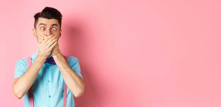 Funny young man covering mouth with hands and squint eyes, making clown grimaces, standing silly on pink background, entertain people