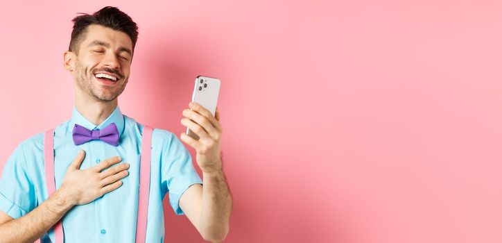 Technology concept. Happy caucasian man video chat and laugh at smartphone, standing over pink background.