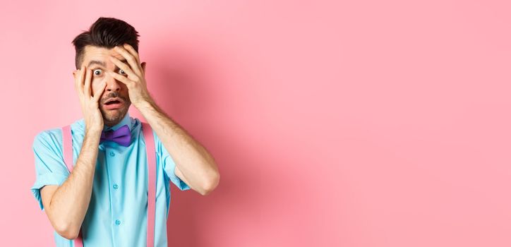 Scared and embarrassed nerdy guy in bow-tie covering his eyes, peeking through fingers at something scary, standing on pink background