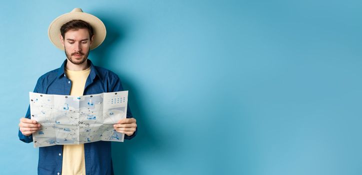 Handsome tourist with summer hat reading map, looking for sightseeing on vacation, standing on blue background