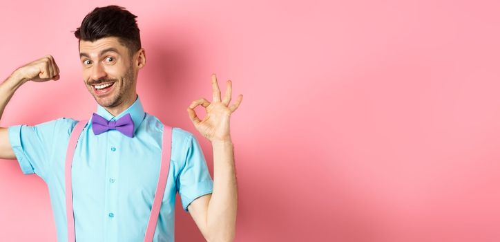 Handsome young man with moustache and bow-tie, flex biceps, showing strong arm and okay sign, workout at gym, standing on pink background