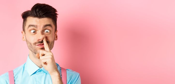 Image of funny caucasian guy picking nose and squint eyes, making dumb face, standing on pink background