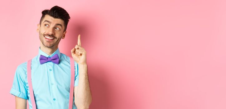 Handsome smiling man raising finger in eureka gesture, looking away and pitching an idea, have suggestion or plan, standing in bow-tie over pink background