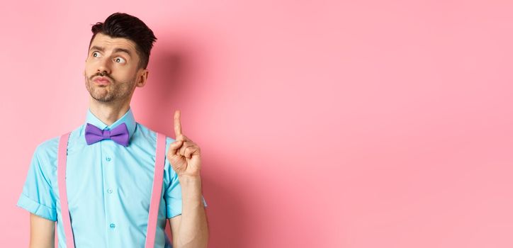 Pensive handsome man in bow-tie pitching an idea, raising index finger while looking away with thoughtful face, standing on pink background