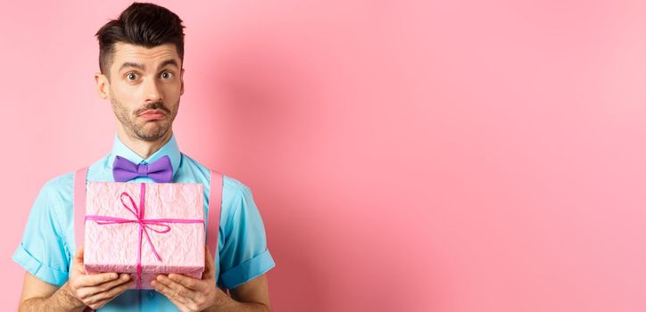 Holidays and celebration concept. Attractive young man with moustache, wearing festive outfit with bow-tie, showing cute gift box and looking at camera, standing over pink background
