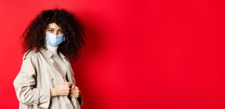 Covid-19, pandemic and quarantine concept. Sassy girl in trench coat and medical mask, put on trench coat for spring walk, red background
