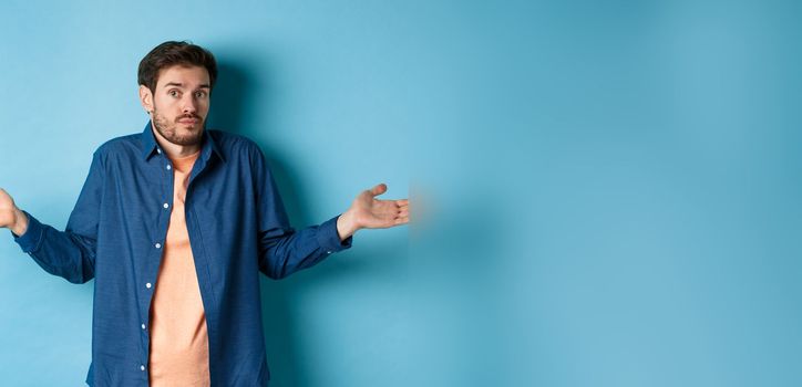 Confused young man with beard, shrugging shoulders and looking clueless at camera, know nothing, standing on blue background
