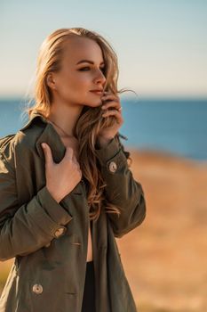 Portrait blonde sea cape. A calm young blonde in an unbuttoned khaki raincoat stands on the seashore, under the raincoat there is a black skirt and top