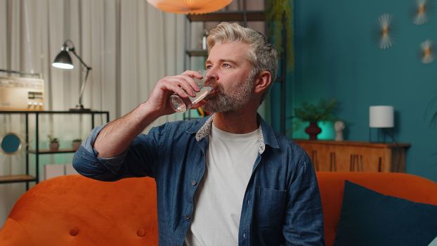 Thirsty middle-aged man sitting at home holding glass of natural aqua make sips drinking still water