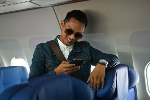 Smiling hipster man traveller in jean jacket leaning on the airline seats and using smart phone