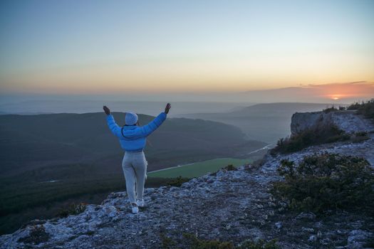 Woman hiker open arms on top of sunrise mountain. The girl salutes the sun, wearing a blue jacket, white hat and white jeans. Conceptual design.