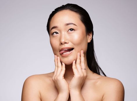 Asian woman, hands and face in beauty skincare for facial cosmetics, botox or treatment against a studio background. Japanese woman smiling with tongue out in satisfaction for hygiene or perfect skin