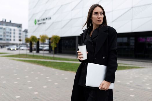 half-length portrait of a stylish businesswoman with a laptop and a glass of coffee against the background of the facade of a modern office building