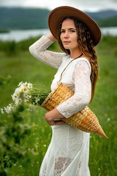 A middle-aged woman in a white dress and brown hat stands on a green field and holds a basket in her hands with a large bouquet of daisies. In the background there are mountains and a lake.