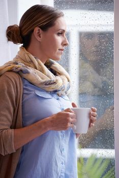Staying warm on a winters day. A young woman gazing out of her window while holding a mug of coffee.