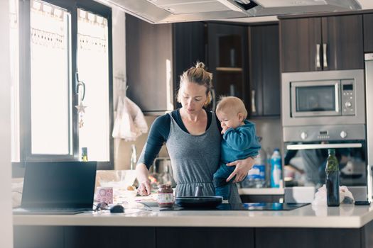 Happy mother and little infant baby boy together making pancakes for breakfast in domestic kitchen. Family, lifestyle, domestic life, food, healthy eating and people concept.
