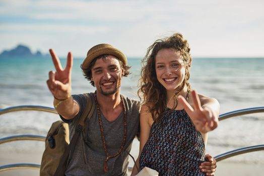 Chilled vibes only. Portrait of a happy young couple showing peace signs while relaxing on a bench by the beach.