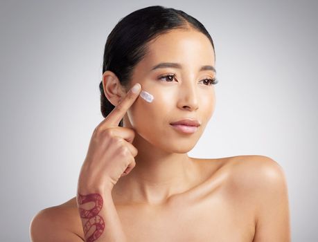 A beautiful smiling mixed race woman applying cream to her cheek. Hispanic model with glowing skin using sunscreen against a grey copyspace background