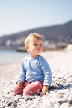 Pensive baby sits, turning to the right, on a pebble beach by the sea against a background of mountains