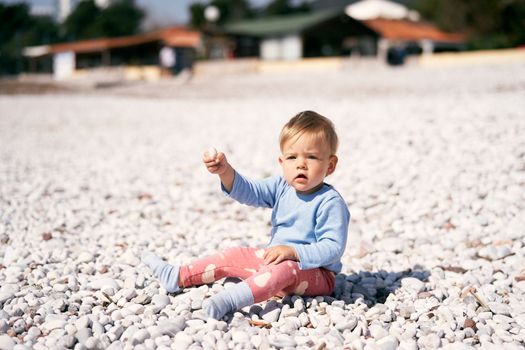 Cute child in a blue blouse and red pants sits on a pebble beach, holding up a pebble in his hand