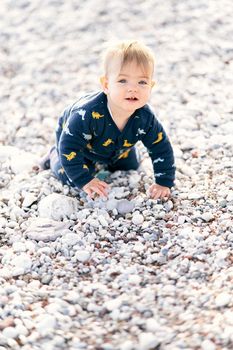 Cute little baby in overalls crawls on a pebble beach