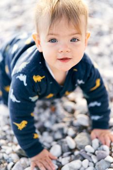 Smiling little baby in overalls sits on a pebble beach. Close-up. Portrait