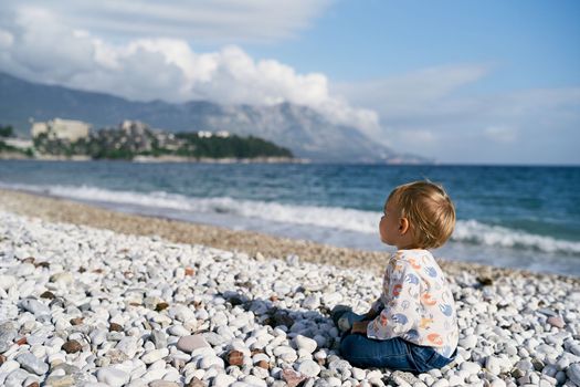 Kid sits on a pebble beach by the sea. Side view