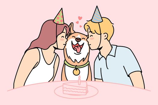 Girl, guy sit at festive table with cake, kiss dog.