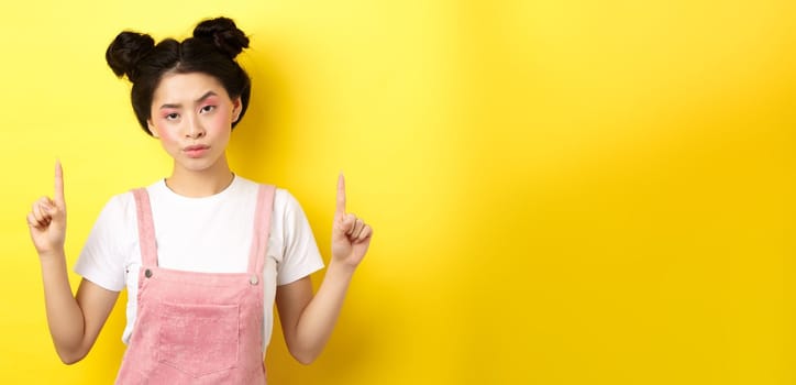 Annoyed and tired asian girl with pink glamour makeup, pointing fingers up and look bothered, standing on yellow background