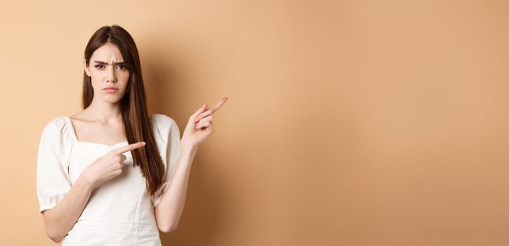 Angry young woman frowning, pointing fingers right and complaining, disappointed with logo, standing upset on beige background
