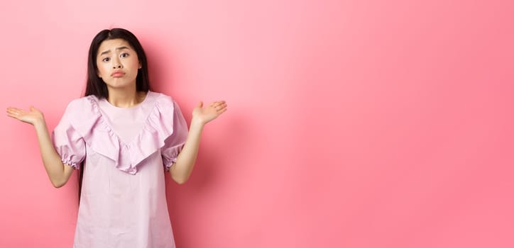 Sorry cant help. Clueless asian girl shrugging with sad face, know nothing, standing indecisive against pink background